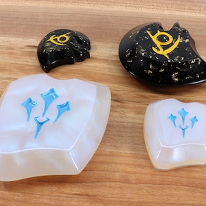May include: Four resin keycaps with different designs. Two keycaps are white with a blue design of three stylized stars. The other two keycaps are black with gold flecks and a gold design of a stylized symbol.