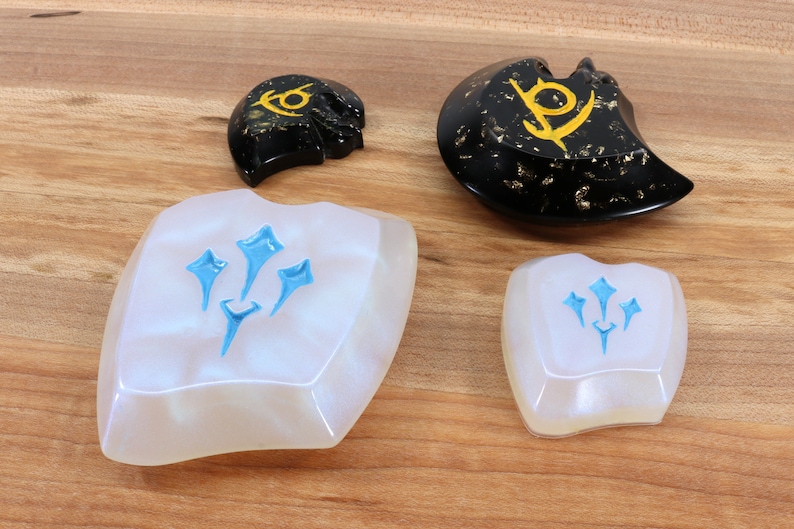 May include: Four resin keycaps with different designs. Two keycaps are white with a blue design of three stylized stars. The other two keycaps are black with gold flecks and a gold design of a stylized symbol.