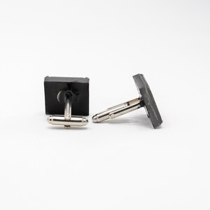 May include: A pair of black cufflinks with silver metal clasps. The cufflinks are square shaped and have a smooth, matte finish.
