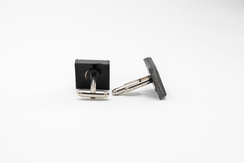May include: A pair of black cufflinks with silver metal clasps. The cufflinks are square shaped and have a smooth, matte finish.