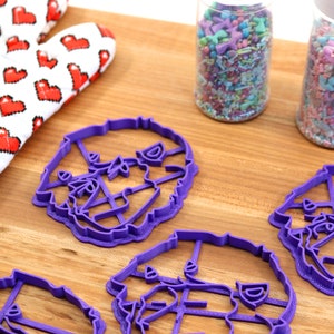May include: Four purple plastic cookie cutters shaped like a dog's head. The cutters are on a wooden surface next to two jars of colorful sprinkles.