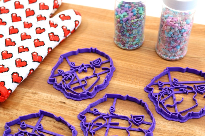 May include: Four purple plastic cookie cutters shaped like a dog's head. The cutters are on a wooden surface next to two jars of colorful sprinkles.