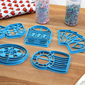 May include: A set of six blue plastic cookie cutters shaped like dice, a slot machine, playing cards, a roulette wheel, and a stack of poker chips. The cutters are on a wooden surface.