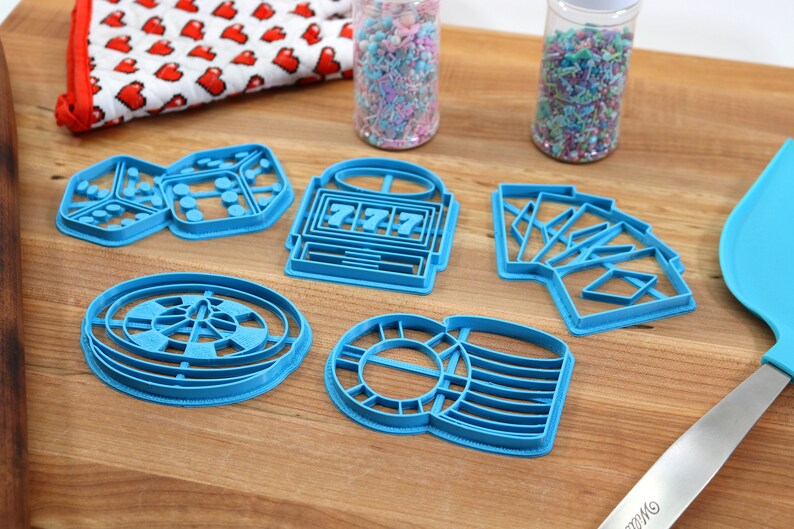 May include: A set of six blue plastic cookie cutters shaped like dice, a slot machine, playing cards, a roulette wheel, and a stack of poker chips. The cutters are on a wooden surface.