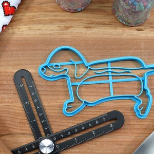 May include: Blue plastic cookie cutter shaped like a dachshund dog. The cutter has a grid pattern inside the body of the dog.