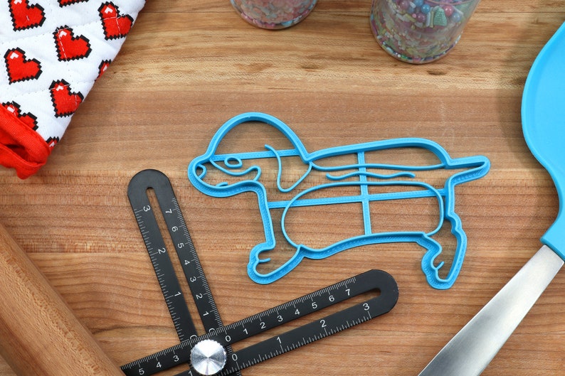 May include: Blue plastic cookie cutter shaped like a dachshund dog. The cutter has a grid pattern inside the body of the dog.