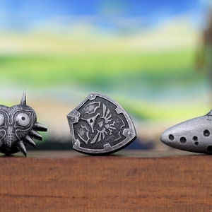 May include: Three silver-coloured metal figurines depicting iconic items from the Legend of Zelda video game series. The figurines are a mask, a shield, and an ocarina.
