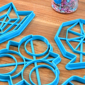 May include: A set of six blue plastic cookie cutters in various geometric shapes, including a star, a circle, a hexagon, and a diamond. The cutters are on a wooden surface.