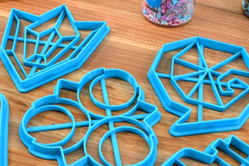 May include: A set of six blue plastic cookie cutters in various geometric shapes, including a star, a circle, a hexagon, and a diamond. The cutters are on a wooden surface.