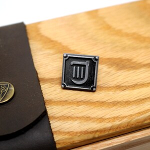 Op de afbeelding: Een zilveren metalen pin met een zwart vierkant frame en een zilveren liersymbool erin. De pin is op een houten oppervlak.