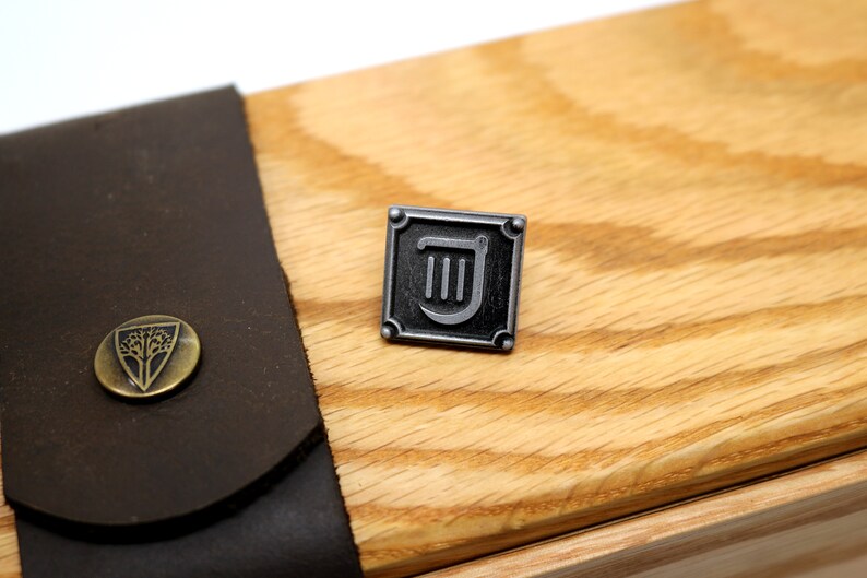 Op de afbeelding: Een zilveren metalen pin met een zwart vierkant frame en een zilveren liersymbool erin. De pin is op een houten oppervlak.