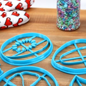 May include: A set of five blue plastic cookie cutters in various shapes, including a circle, an oval, and a leaf. The cutters are on a wooden surface.