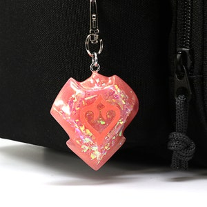 May include: A pink iridescent charm with a stylised heart and crown design, attached to a silver keychain.