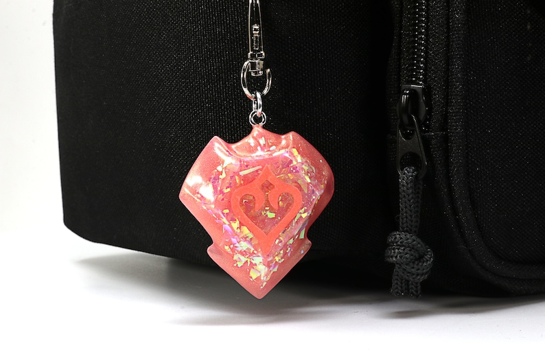 May include: A pink iridescent charm with a stylised heart and crown design, attached to a silver keychain.