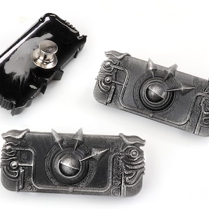 May include: Three metal pins, two with a detailed design and one with a black backing. The pins are shaped like a rectangle with a circular design in the centre. The pins have a silver finish.