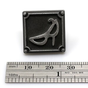 May include: A silver metal button with a raised design of a high-heeled shoe. The button is square with rounded corners and has four small decorative dots on the corners.