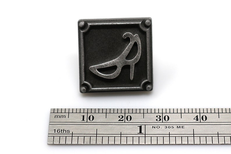 May include: A silver metal button with a raised design of a high-heeled shoe. The button is square with rounded corners and has four small decorative dots on the corners.