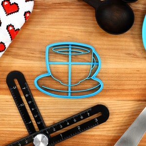 May include: A blue plastic coffee cup-shaped cookie cutter sits on a wooden surface, next to a heart-patterned oven mitt, a black angle finder, a blue spatula, and black measuring spoons. Baking and kitchen tools.