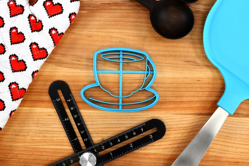 May include: A blue plastic coffee cup-shaped cookie cutter sits on a wooden surface, next to a heart-patterned oven mitt, a black angle finder, a blue spatula, and black measuring spoons. Baking and kitchen tools.