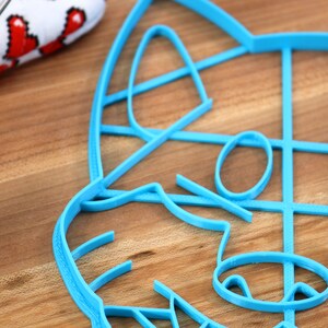 May include: A blue plastic cookie cutter shaped like a raccoon face. The cutter is on a wooden surface.