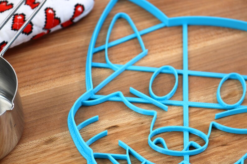 May include: A blue plastic cookie cutter shaped like a raccoon face. The cutter is on a wooden surface.