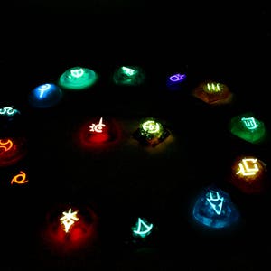 May include: A collection of 14 glowing, colorful, round, resin dice with various symbols on each face. The dice are arranged in a circle on a dark background.