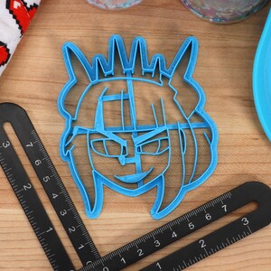 May include: A blue plastic cookie cutter shaped like a cartoon character with a crown, horns, and a mischievous expression.