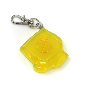 May include: A translucent yellow keychain with a silver clasp. The keychain has a square shape with a curved bottom edge and an orange symbol in the center. The symbol appears to be a stylized letter or emblem.