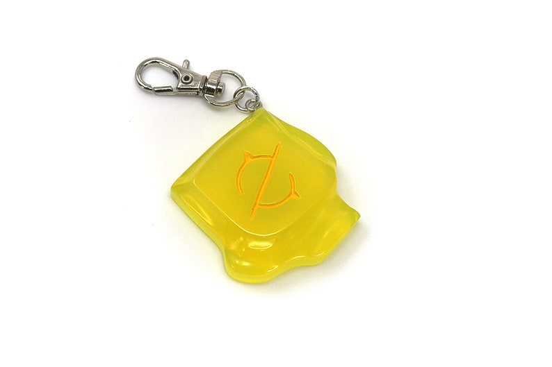 May include: A translucent yellow keychain with a silver clasp. The keychain has a square shape with a curved bottom edge and an orange symbol in the center. The symbol appears to be a stylized letter or emblem.