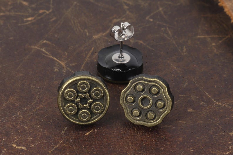 以下が含まれることがあります： アンティークスタイルのスタッドピアス3つ。銃のシリンダーのデザイン。ピアスはブロンズ色の金属と黒い裏地で作られています。1つのピアスは、ポストとバタフライバックが付いています。