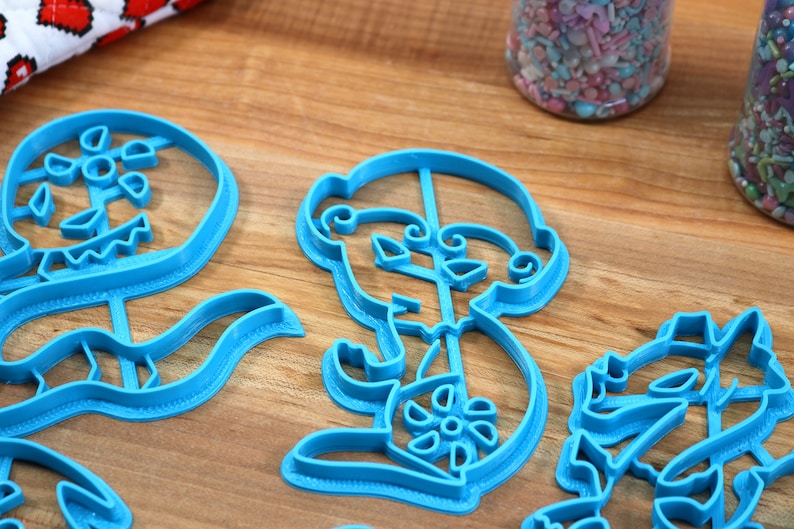 May include: A set of four blue plastic cookie cutters shaped like a skeleton holding a flower, a flower, a skeleton holding a flower, and a flower.
