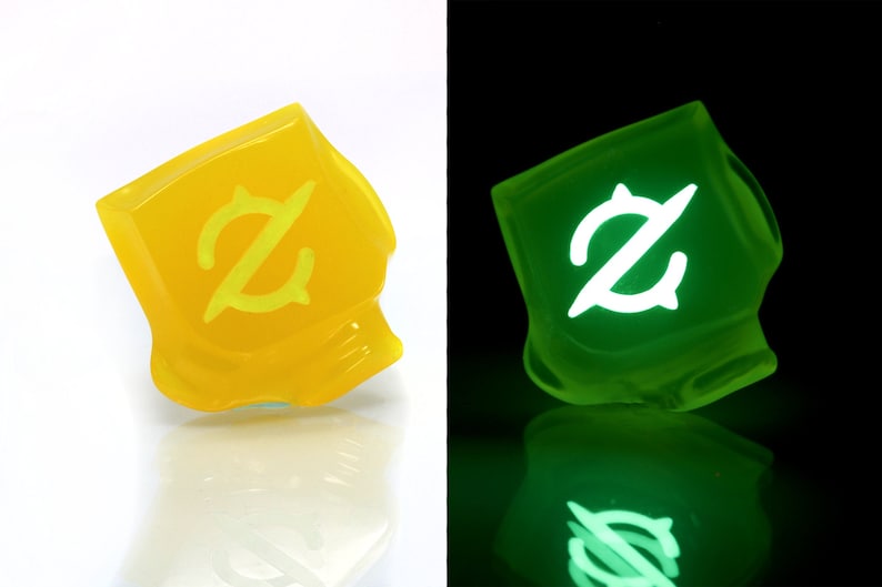 以下が含まれることがあります： スタイリッシュな「Z」デザインの、半透明のオレンジ色の正方形のオブジェクト。暗闇では緑色に光ります。デザインは、各端に円形の要素が付いた様式化された文字「Z」です。背景は白と黒です。