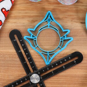 May include: A blue plastic cookie cutter shaped like a stylized sun with a circular center. The cutter is on a wooden surface with a black ruler.