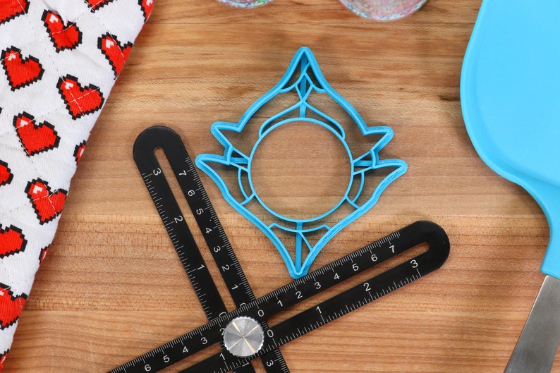 May include: A blue plastic cookie cutter shaped like a stylized sun with a circular center. The cutter is on a wooden surface with a black ruler.