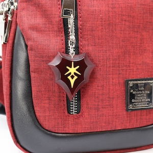 May include: Red crossbody bag with a black zipper and a black leather trim. A silver chain with a red and gold shield charm hangs from the zipper. The charm has a stylized symbol on it. The bag has a small silver tag that reads "Where is the heart to find the good where good is there."