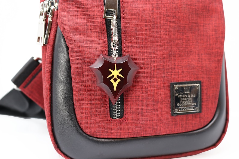 May include: Red crossbody bag with a black zipper and a black leather trim. A silver chain with a red and gold shield charm hangs from the zipper. The charm has a stylized symbol on it. The bag has a small silver tag that reads "Where is the heart to find the good where good is there."