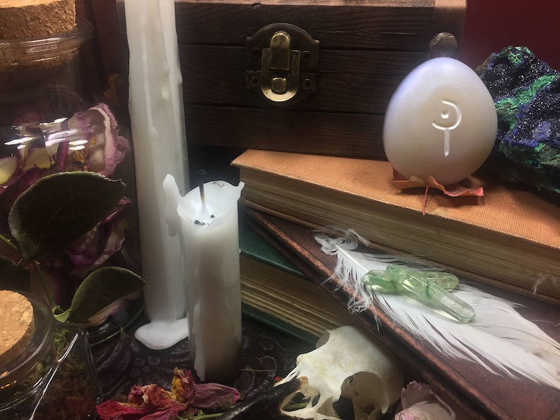 May include: A white candle with a white egg-shaped object with a symbol on it, a wooden box, and a book with a feather and a green crystal on top.