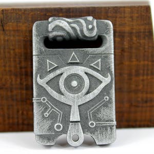 May include: A silver-coloured metal pendant featuring a detailed design of an eye with a triangle above and below it. The pendant has a textured surface and a loop at the top for attaching a chain.