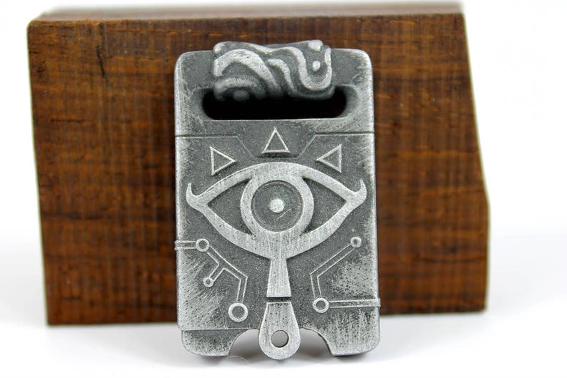 May include: A silver-coloured metal pendant featuring a detailed design of an eye with a triangle above and below it. The pendant has a textured surface and a loop at the top for attaching a chain.