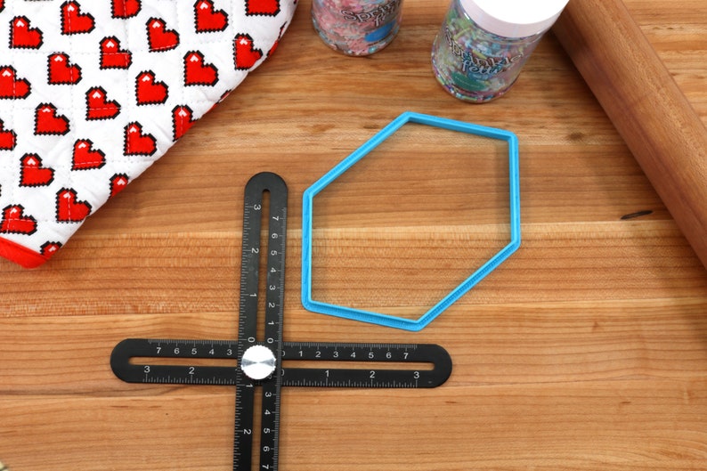 Op de afbeelding: Een blauwe plastic zeshoekige koekjesvormer op een houten oppervlak met een zwarte liniaal en een deegroller.