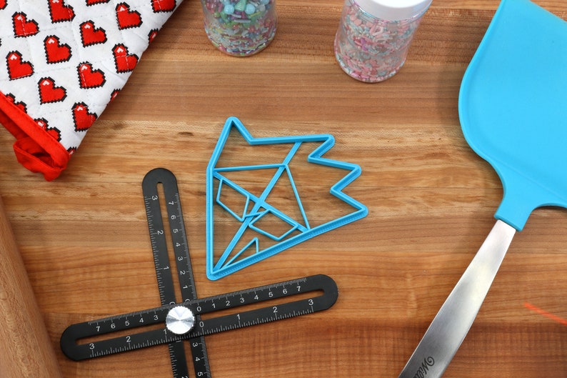 May include: A blue plastic cookie cutter shaped like a geometric star on a wooden surface with a ruler, a spatula, and two jars of sprinkles.