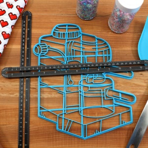 May include: Blue plastic cookie cutter shaped like an espresso machine. The cutter is on a wooden surface with a ruler, a rolling pin, a spatula, and two jars of sprinkles.