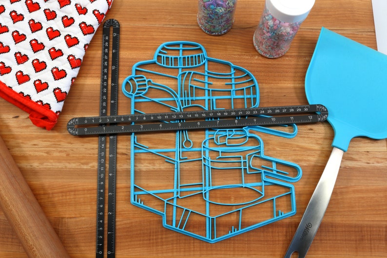 May include: Blue plastic cookie cutter shaped like an espresso machine. The cutter is on a wooden surface with a ruler, a rolling pin, a spatula, and two jars of sprinkles.