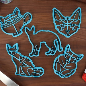 Puede incluir: Conjunto de cinco cortadores de galletas de plástico azul con forma de gato. Los cortadores están en varias poses, incluyendo un gato sentado, un gato de pie, un gato con una taza de té y una cabeza de gato.