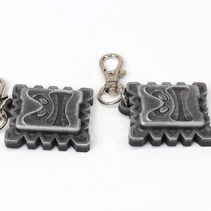May include: Two silver keychains with a detailed design of a face with a geometric pattern. Each keychain has a silver metal loop.