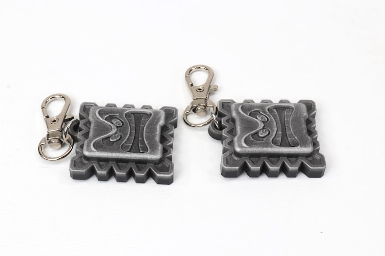 May include: Two silver keychains with a detailed design of a face with a geometric pattern. Each keychain has a silver metal loop.