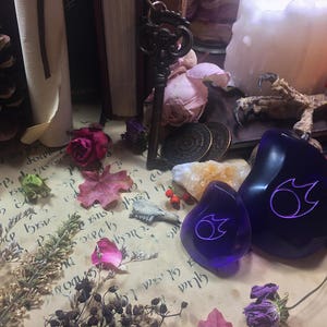 Op de afbeelding: Twee paarse glazen stenen met een wit vlamontwerp. De stenen liggen op een perkament-achtig oppervlak met gedroogde bloemen en andere natuurlijke elementen.