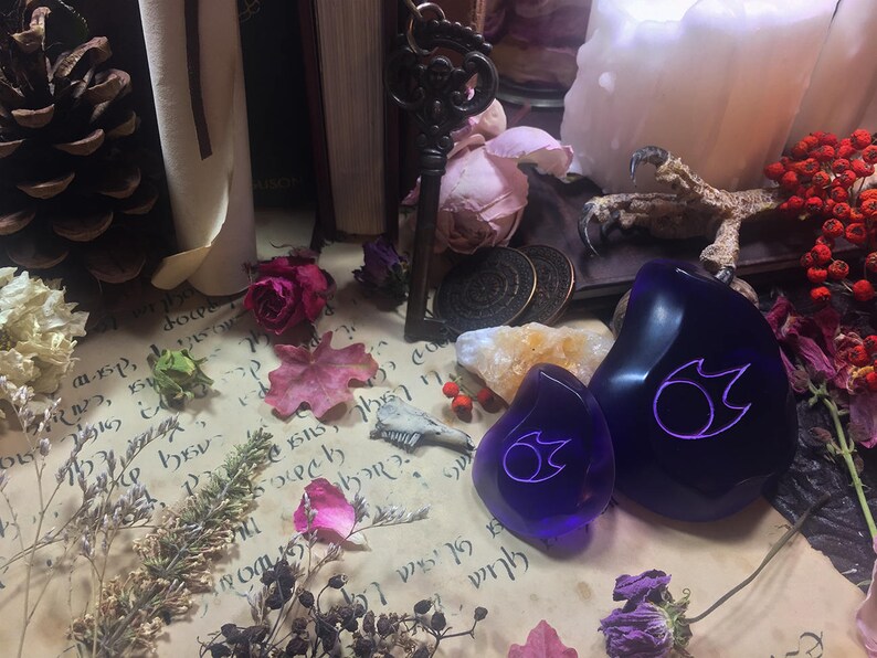 Op de afbeelding: Twee paarse glazen stenen met een wit vlamontwerp. De stenen liggen op een perkament-achtig oppervlak met gedroogde bloemen en andere natuurlijke elementen.