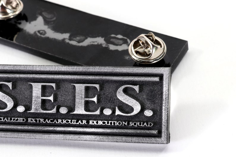 May include: A silver-colored metal pin with the text "S.E.E.S. SPECIALIZED EXTRACURRICULAR EXECUTION SQUAD" on it.