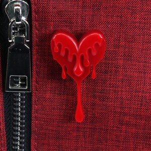 May include: A red heart-shaped pin with dripping red paint. The pin is attached to a red fabric background.